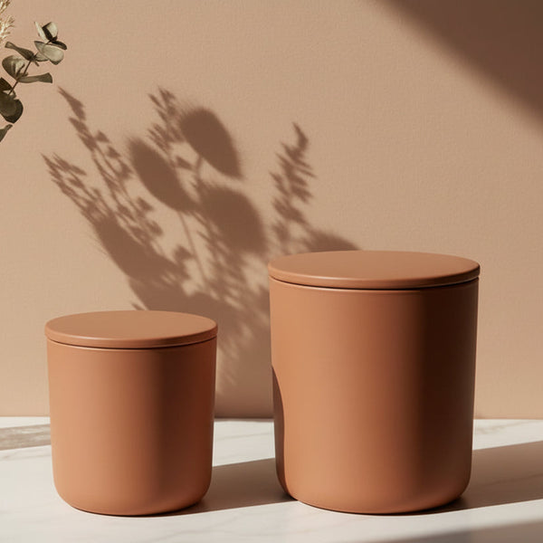 Two terracotta-colored storage jars with lids on a light surface against a beige wall with plant shadows.